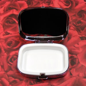 May include: A silver-colored, rectangular trinket box with a hinged lid. The lid is open, revealing a white interior. The box is sitting on a red background.