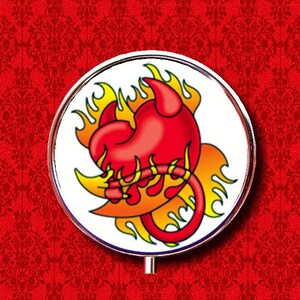 May include: A round, white compact mirror with a red heart with devil horns and a tail, surrounded by yellow flames. The heart is on a white background.