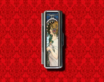 May include: A silver metal case with a hinged lid. The case features a colorful art nouveau design of a woman with a flower in her hair. The case is approximately 2 inches tall and 1 inch wide.
