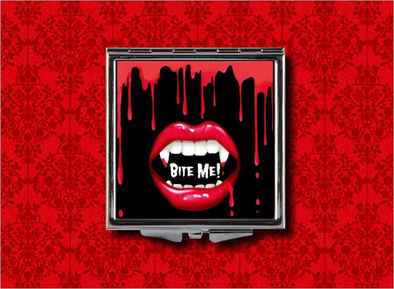 Puede incluir: Un espejo compacto con fondo negro y una boca de vampiro roja con colmillos. La boca gotea sangre roja y el texto "Bite Me!" est&aacute; escrito en letras blancas.