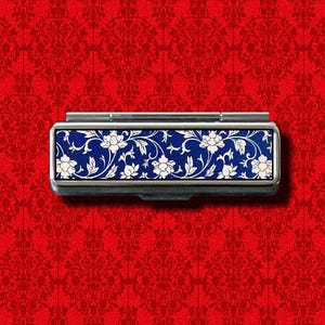 Puede incluir: Una caja de metal plateado con un diseño floral azul y blanco. La caja tiene una tapa con bisagras y es de tamaño pequeño.