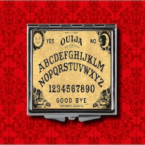 May include: A silver compact mirror with a vintage Ouija board design. The board features the letters A-Z, numbers 0-9, "Yes", "No", "Goodbye", and a sun and moon graphic.