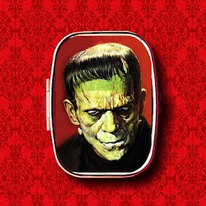 May include: A vintage-style metal tin featuring a green-faced monster with black hair. The tin has a red background and a silver hinge.