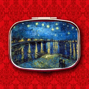 May include: A compact mirror with a silver frame and a painted image of a starry night sky over a river with a town in the distance. The painting is in the style of Vincent van Gogh.