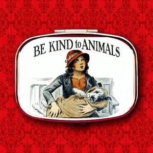 May include: A white metal compact with a red and white image of a girl holding a dog. The compact has the text "BE KIND to ANIMALS" printed on it.