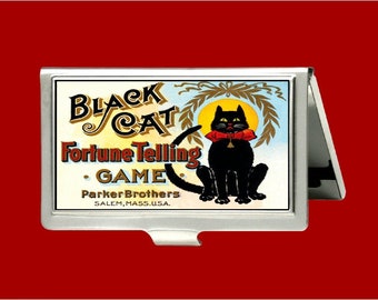 Può includere: Un porta carte in argento con un'illustrazione vintage di un gatto nero con un papillon rosso. L'illustrazione è intitolata "Black Cat Fortune Telling Game" e include il testo "Parker Brothers Salem, Mass. USA."