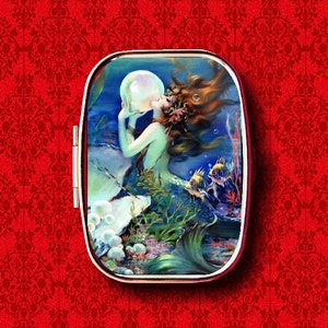 May include: A vintage-style metal compact with a colorful image of a mermaid holding a pearl. The mermaid has long flowing red hair and a green tail. The background is a blue ocean with bubbles and fish.