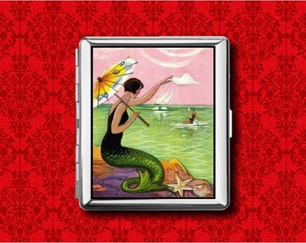 May include: A silver cigarette case featuring a vintage illustration of a mermaid on a rocky shore. The mermaid, with a green tail and black swimsuit, holds a parasol and reaches towards a sailboat. The background is a pink sky and green water.