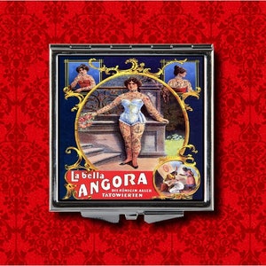 Pu&ograve; includere: Uno specchio compatto vintage con sfondo blu e cornice dorata. Lo specchio presenta un'immagine in bianco e nero di una donna con tatuaggi, con il testo "La bella Angora Die K&ouml;nigin aller T&auml;towierten" in tedesco.