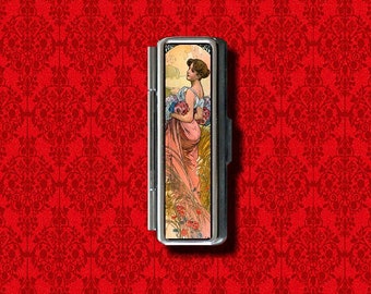 Puede incluir: Un estuche compacto de metal plateado con una imagen colorida de una mujer en un campo de flores. La imagen es de estilo Art Nouveau.