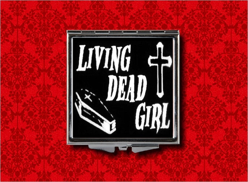 May include: A black and white compact mirror with the text "Living Dead Girl" and a white coffin with a cross on it. There is also a white cross on the right side of the mirror.