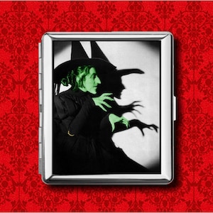 May include: A silver cigarette case featuring a black and white image of the Wicked Witch of the West from The Wizard of Oz. The witch is depicted with green skin and a black hat, casting a long shadow. The background is a red floral pattern.