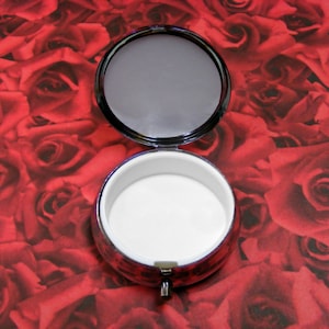 May include: A small, round, silver-colored trinket box with a white interior. The box is open and the lid is laying on top of the box. The box is sitting on a red background with a pattern of roses.