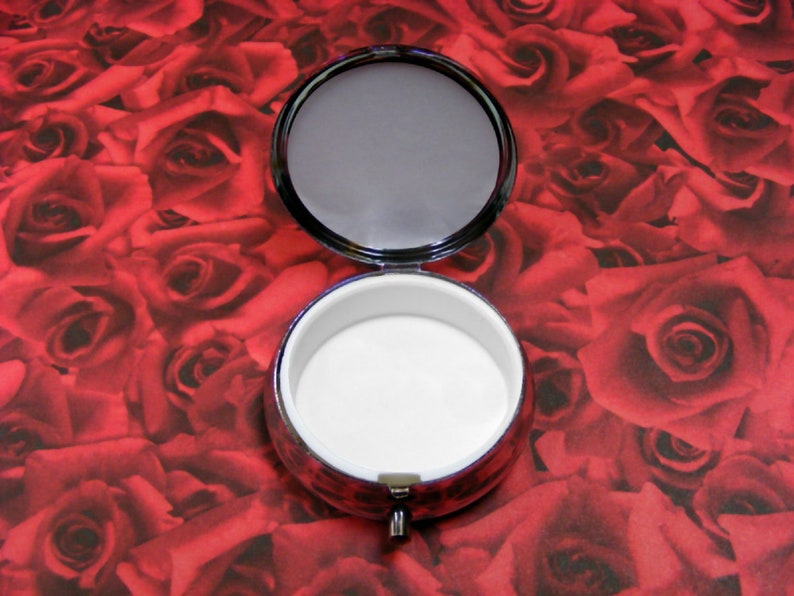 May include: A small, round, silver-colored trinket box with a white interior. The box is open and the lid is laying on top of the box. The box is sitting on a red background with a pattern of roses.