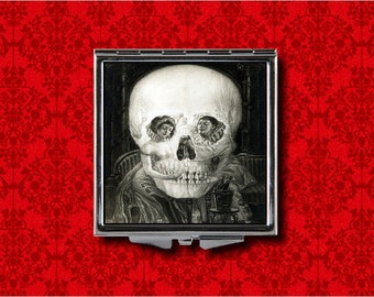 May include: A silver compact mirror with a black and white image of a skull with two people's faces inside. The image is a classic optical illusion.