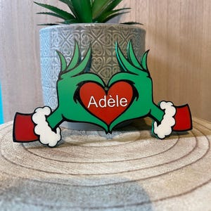 Peut inclure: Un ornement décoratif représentant deux mains vertes aux longs doigts formant un cœur autour d'un cœur rouge. Le cœur rouge porte le prénom Adèle écrit en blanc. Les mains ont des manches rouges avec des poignets en fourrure blanche.