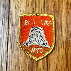 May include: A red and gold embroidered patch with the words "Devils Tower" and "WYO" in white. The patch features a white embroidered image of Devils Tower National Monument in Wyoming.