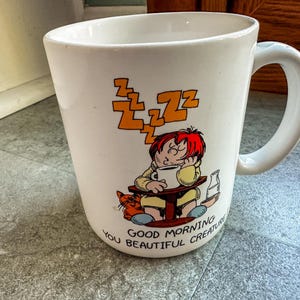 May include: White ceramic mug with a cartoon illustration of a person sleeping in a chair with a cat and the text "Good Morning You Beautiful Creature".
