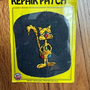 May include: A yellow cat with red, green, and blue accents on a black fabric patch. The patch is labeled "Repair Patch" and "Made, Packaged, Printed in U.S.A."