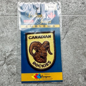 May include: A yellow and brown embroidered patch with the text "Canadian Rockies" and a brown mountain goat illustration. The patch is on a blue background with the text "Collector Series" and "Canada Souvenirs" in white.