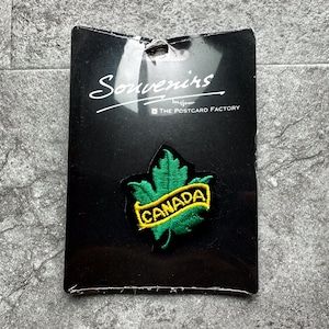 May include: A black card with the words "Souvenirs" and "The Postcard Factory" printed on it. A green felt maple leaf with a yellow felt banner that reads "CANADA" is attached to the card.