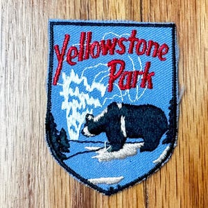 May include: Blue embroidered patch with the text "Yellowstone Park" in red lettering. A black bear with white markings is standing in front of a white geyser.