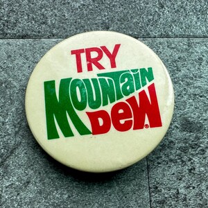 May include: A round, off-white button with the words "TRY MOUNTAIN DEW" in red and green lettering. The button is a vintage promotional item, likely from a soft drink campaign.