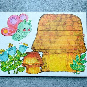 May include: A rectangular stationery sheet featuring a large, golden-brown mushroom with horizontal lines for writing. To the left, a light green caterpillar with pink and purple butterfly wings sits above three blue bell-shaped flowers and a smaller mushroom.