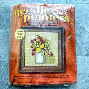 May include: Needlepoint kit featuring a design of a white ceramic pot with a bouquet of yellow and red daisies. The kit includes printed Aida cloth, 100% wool yarn, needles, a frame offer inside, and complete instructions. The kit is from Needlepointers, 2400 Olive Ave, Concord, California 94520.