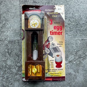 May include: A vintage egg timer shaped like a miniature grandfather clock. The brown plastic clock has a gold-colored face and a pendulum. The packaging reads "3 minute egg timer" with a graphic of a telephone and an egg.
