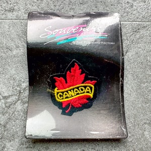 May include: A red and yellow embroidered patch with a maple leaf and the word "CANADA" on a black background. The patch is packaged on a black card with the text "Souvenirs The Postcard Factory".
