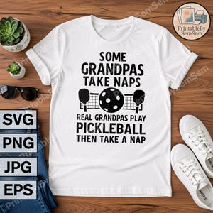 May include: White t-shirt with the text "SOME GRANDPAS TAKE NAPS, REAL GRANDPAS PLAY PICKLEBALL THEN TAKE A NAP" in black. The design includes pickleball paddles, a ball, and a net. The shirt is displayed on a wooden surface.