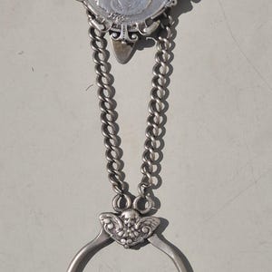 May include: An antique silver coin holder with a ring handle. The coin is set in an ornate frame and connected to the ring by two chains. The ring features a decorative skull and wing design. The coin holder is likely from the late 1800s or early 1900s.