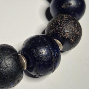 May include: Close-up of a strand of dark blue, spherical beads. The beads have a textured surface and are separated by small, silver-colored spacers. The beads appear to be made of glass or a similar material.