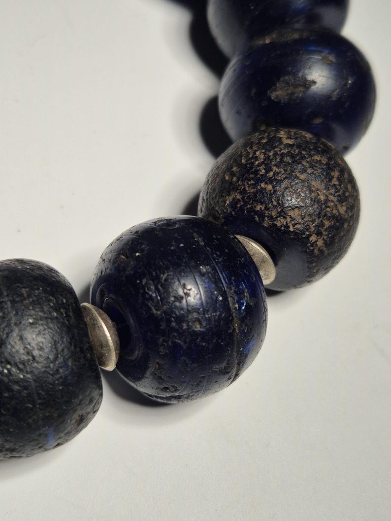 May include: Close-up of a strand of dark blue, spherical beads. The beads have a textured surface and are separated by small, silver-colored spacers. The beads appear to be made of glass or a similar material.