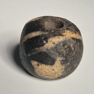 May include: A round, dark-colored stone with a hole on top. The stone features a pattern of black and tan stripes. The surface appears textured and worn, suggesting age or natural weathering. The object is likely a decorative or collectible item.