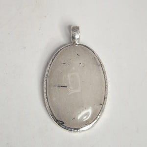 Könnte beinhalten: Ovaler Anhänger mit einem klaren, hellgrauen Stein, der dunkle Einschlüsse und einen silberfarbenen Metallrahmen aufweist. Der Anhänger hat oben eine Schlaufe für eine Kette. Der Stein hat eine rechteckige Form.