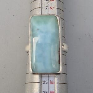 May include: A rectangular silver ring featuring a large, light blue gemstone with a marbled appearance. The ring is set on a ring sizer, showing size measurements. The gemstone has a light blue and white color.