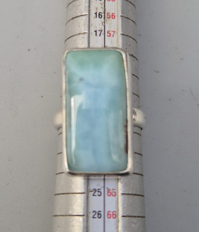 May include: A rectangular silver ring featuring a large, light blue gemstone with a marbled appearance. The ring is set on a ring sizer, showing size measurements. The gemstone has a light blue and white color.