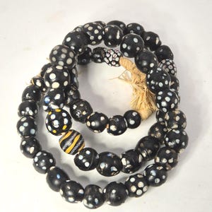 Könnte beinhalten: Drei Stränge schwarzer Perlen mit weißen Tupfen und ein paar gelb-schwarz gestreiften Perlen. Die Perlen sind aufgefädelt und mit Naturfaser gebunden. Die Perlen werden wahrscheinlich für Schmuck oder Dekorationszwecke verwendet.