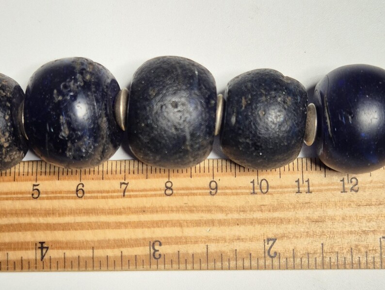May include: Close-up of a strand of large, dark blue beads with silver-colored spacers. The beads are roughly 1 inch in diameter. A wooden ruler with inch markings is in the foreground, providing scale.