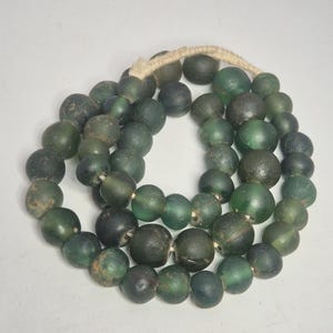 Könnte beinhalten: Eine Halskette aus dunkelgrünen Glasperlen. Die Perlen variieren in Größe und Form und sind an einer Kordel aus Naturfaser aufgereiht. Die Perlen haben ein leicht unregelmäßiges, gealtertes Aussehen mit Farb- und Texturvariationen.