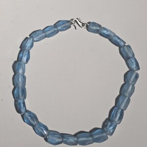 May include: A necklace composed of translucent, light blue, faceted beads arranged in a circular pattern. The beads have a matte, frosted texture and are connected by a silver-toned metal S-hook clasp at the top, securing the strand together.