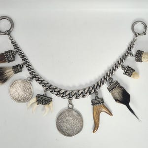 Könnte beinhalten: Ein silberfarbenes Charm-Armband mit verschiedenen Anhängern, darunter Tierkrallen, Fell, Hörner und Münzen. Das Armband hat ein Panzerketten-Design mit einem Verschluss an jedem Ende. Die Charms sind mit dekorativen silberfarbenen Kappen befestigt.