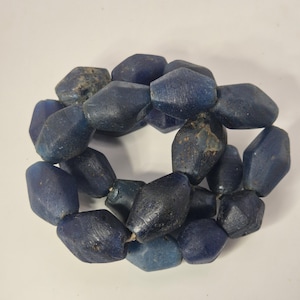 Könnte beinhalten: Eine Kette aus dunkelblauen, facettierten Perlen. Die Perlen haben eine unregelmäßige, geometrische Form und eine leicht raue Textur. Die Perlen sind aufgefädelt und bilden eine kreisförmige Form. Die Farbe der Perlen variiert leicht.