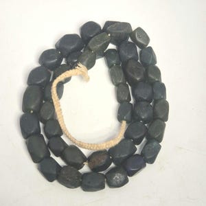 Könnte beinhalten: Eine Halskette aus dunklen, unregelmäßig geformten Perlen. Die Perlen sind auf einer hellen, gewebten Kordel aufgefädelt. Die Perlen haben eine dunkle, fast schwarze Farbe mit einigen Farbvariationen.