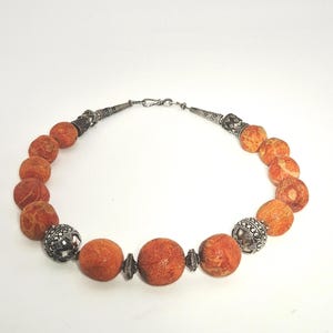 Può includere: Una collana con grandi perle rotonde arancioni, intervallate da perle e dettagli ornamentali in metallo color argento. La collana è infilata su una catena e una chiusura color argento, creando un contrasto sorprendente.