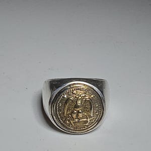 Puede incluir: Anillo de sello plateado con un emblema dorado con un diseño de águila. El anillo tiene una banda ancha y una cara circular con detalles en relieve. Una pieza de joyería clásica.