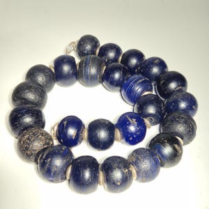 May include: A necklace of dark blue and black beads. The beads are round and have a slightly rough texture. Some beads have a striped pattern. The necklace is strung on a thin, light-colored cord.