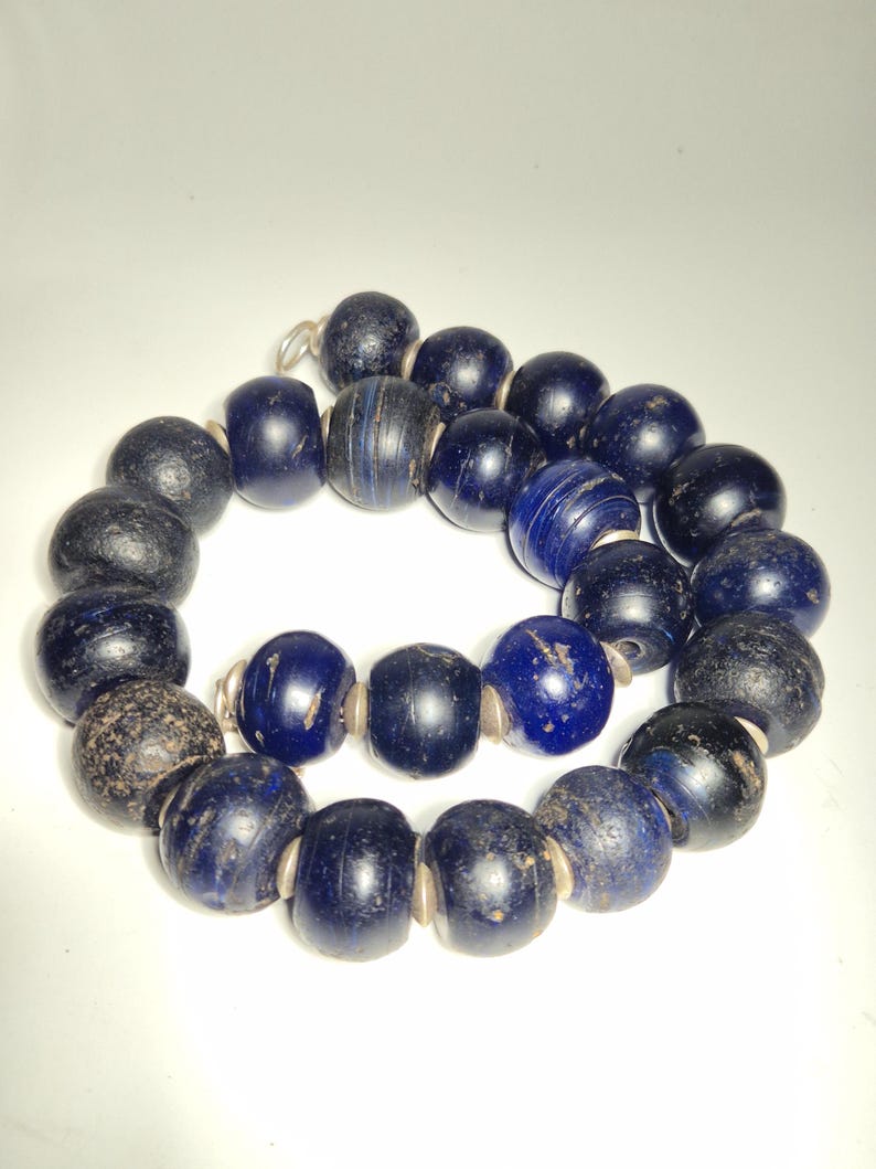 May include: A necklace of dark blue and black beads. The beads are round and have a slightly rough texture. Some beads have a striped pattern. The necklace is strung on a thin, light-colored cord.